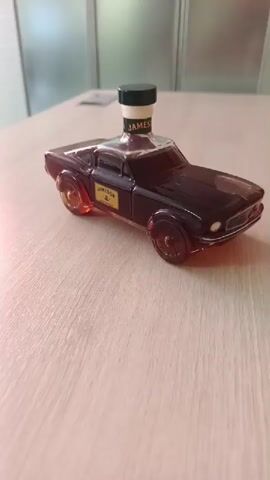 A miniature bottle of Jameson Irish Whiskey, designed to resemble a classic muscle car, sits on a light brown wooden table. The bottle is dark brown, shaped like a car, with a clear bottle cap on top. A yellow label with black text is visible on the side of the car-shaped bottle. The background shows a lightly colored wall with a window on the left, and a blurred office chair is visible in the upper left corner. The lighting appears to be artificial, casting a warm glow on the scene. The camera pans slightly, focusing on the bottle.