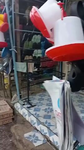The video shows a bustling street market on a cloudy day. The street is wet and appears slightly muddy. Stalls line the side of the street, selling various goods. The stall closest to the camera displays stacks of cardboard boxes and rows of shoes and sandals on shelves. Hanging above are plastic bird feeders in yellow, red and white. Motorcycles carrying passengers weave through the crowd. People are walking and standing around, browsing the market. A white van is parked on the left side of the street. The buildings are simple, some with corrugated metal roofs.