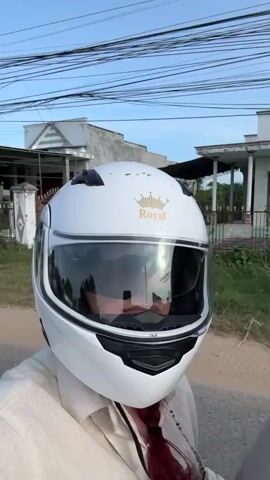 The video shows two people riding a motorcycle down a road lined with trees and houses. The person in the foreground is wearing a white helmet with a gold crown logo and a tan jacket. The person behind them is wearing a black helmet and a grey shirt. The sky is cloudy with the sun shining through. The road is paved and appears to be in a rural area.