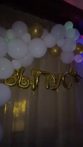This video captures a celebratory event, likely a graduation party or similar milestone, given the large "ВЫПУСК" (Graduation) balloon sign. The scene is set indoors, with a dimly lit atmosphere enhanced by colorful disco lights projected onto the ceiling. White and gold balloons form a decorative arch, adding to the festive mood.

In the foreground, a cluster of white balloons is visible, some reflecting the ambient light. Behind them, the prominent gold balloon letters spell out "ВЫПУСК". The background reveals a gathering of people dancing and socializing. The presence of multiple individuals enjoying themselves suggests a lively party. The disco lights, with their shifting patterns of blue, pink, green, and yellow, create a dynamic and energetic ambiance. The overall impression is one of joy and celebration, with the event taking place at night, indicated by the darkness outside the venue and the artificial lighting within. The setting appears to be a hall or event space, with curtains and wooden beams visible.