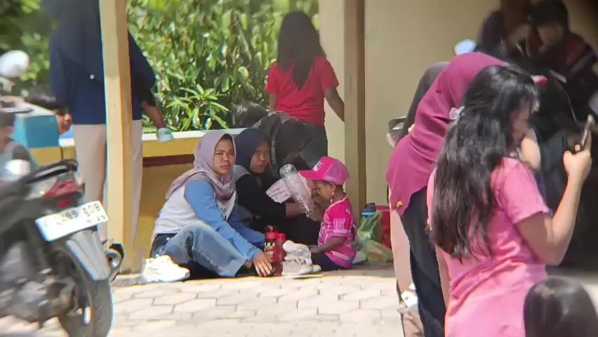 The video shows a group of people, mostly women and children, sitting and standing outside a building. In the foreground, a motorbike is parked. Some individuals are wearing headscarves. One woman in a pink shirt is holding a phone to her ear. Another woman in a purple headscarf is talking to someone. A few children are visible, one wearing a pink cap. The background shows greenery and part of a building with a yellow wall. The ground is paved with interlocking bricks.