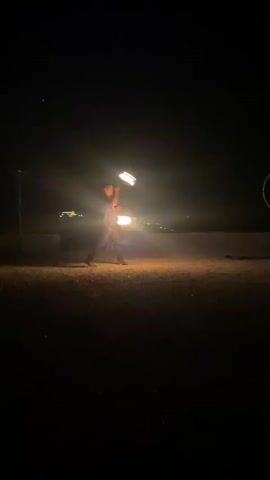 A person is performing with fire in front of a dark background. They are holding two flaming objects and appear to be juggling them. There are lights in the distance behind the performer. The ground is dark and appears to be dirt or sand. The video also briefly shows a red heart-shaped light and then a distant view of buildings with lights.