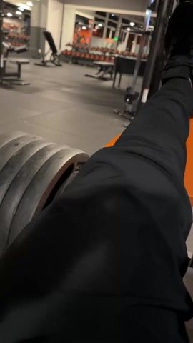 A person is performing leg presses in a gym. The camera is positioned near their leg, showing their black-clad leg extended towards a leg press machine loaded with weights. The gym has a gray and orange color scheme and is equipped with various fitness machines. The person's foot is visible, pressing against the machine.
