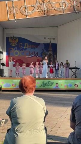 An outdoor stage is set with a backdrop featuring the Russian flag colors, the text "С Днем России" (Happy Russia Day), "12 ИЮНЯ" (12 JUNE), the Russian coat of arms, and St. Basil's Cathedral. A woman in a light blue jumpsuit sings into a microphone at the center of the stage. To her left, five young girls in matching pink and white polka dot dresses clap their hands. Further left, another woman in a white dress stands beside a speaker. To the right of the singer, three children stand: a boy in a red vest, a boy in a plaid vest, and a girl in a white and floral dress holding a bouquet. The front of the stage is decorated with a banner displaying "Лето" (Summer). In the foreground, from behind, an audience member with red hair in a light green top watches the performance, with another person partially visible to her right. The scene is brightly lit, suggesting an outdoor daytime event.