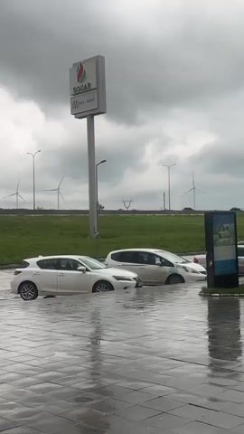 The video shows two white cars partially submerged in standing water on a paved surface. The water level reaches the lower portion of the car doors. A sign for SOCAR and WAY MART is visible in the background. Beyond the sign, there is a green field with wind turbines and a highway with moving vehicles. The sky is overcast with dark clouds. Rain has likely occurred recently.
