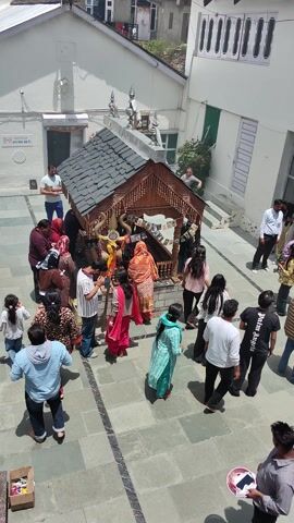 The video captures a lively scene outside a temple in Kishorl, India. The primary subjects are a diverse group of people, including men, women, and children, gathered in a courtyard paved with flagstones. Some are dressed in traditional Indian attire, such as colorful sarees and kurtas, while others wear more contemporary clothing.

The scene is set against the backdrop of a temple complex. A prominent white dome structure with gold ornamentation is visible to the left, likely a central part of the temple. To the right, a smaller, intricately carved wooden structure, possibly a shrine or a decorative element, is laden with offerings and surrounded by people. Buildings with multiple windows and balconies are also visible, suggesting a populated area or adjacent structures.

The people appear to be engaged in various activities, primarily related to a temple visit or a religious gathering. Some are standing in a line, seemingly waiting to enter a structure or receive blessings. Others are interacting with each other, and some are observing the surroundings. The overall mood seems to be one of reverence and community engagement.

Notable details include the bright daylight, suggesting it's daytime, and the presence of fluffy white clouds against a blue sky, indicating fair weather. The architecture, the attire of the people, and the mountainous landscape visible in the distance all point towards a North Indian or Himalayan region, consistent with the provided location context.

Visible text includes signage in a South Asian script on one of the white buildings. While the text is not clearly legible due to the image quality and angle, it adds to the cultural context of the location. The overall impression is a vibrant cultural and religious event taking place in a picturesque, mountainous setting.