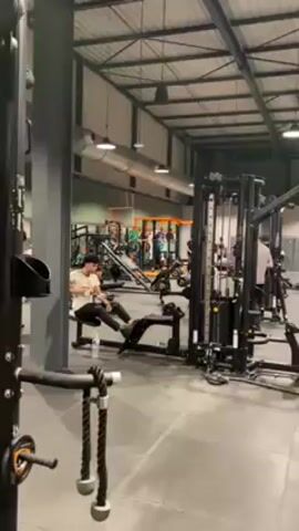 The video shows the interior of a gym. The gym has a high ceiling with exposed metal beams and pipes. The floor is a dark grey tiled surface. There is exercise equipment scattered throughout, including cable machines, rowing machines, and weight benches. There are people exercising in the gym. A person is seen using a leg curl machine in the foreground. There are others in the background moving around and using other equipment. The overall lighting in the gym is bright.