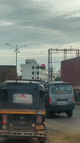 The video shows a street scene with several vehicles. In the foreground, the rear of a black and yellow auto-rickshaw is visible. It has a sign on the back that appears to be in a script other than English. Behind the auto-rickshaw, a large brown truck is visible, and to the right, the side of a grey van is seen. The sky is overcast with clouds. A street light with multiple arms is also visible. A small cluster of colorful balloons is attached to a pole between the auto-rickshaw and the truck. The vehicles are stationary, suggesting traffic.