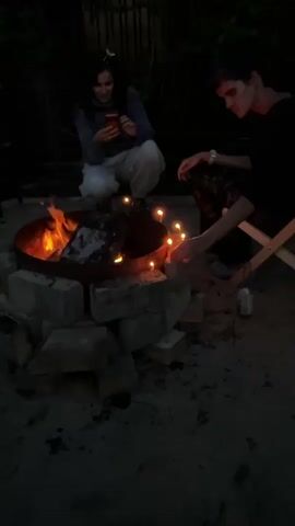 Two individuals are present outdoors in a dimly lit environment, likely at night, around a fire pit. The fire pit is constructed from bricks. In the center of the fire pit, burning logs are visible, emitting flames and light.

To the left, a person with dark, long hair is crouched down, looking at a red smartphone. They are wearing a blue-gray hooded sweatshirt and light-colored pants.

To the right, another individual with short, dark hair is also crouched. Their hands are near the fire pit, one hand extended towards the flames and the other resting near a small, yellow, rectangular object that appears to be a cake with lit candles. This person is wearing dark clothing with a patterned design. A white folding chair is visible behind this individual.

In the background, a dark fence or structure is partially visible. The ground surface appears to be dirt or sand, with scattered debris. The overall ambiance suggests a casual outdoor gathering, possibly a birthday celebration, given the presence of the cake and candles. A small bottle is also visible on the ground to the right of the cake.