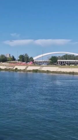 A river flows under a clear blue sky, with a "ONLY LYON" sign visible on the opposite bank. The camera pans left, revealing a modern, geometrically-shaped building on the riverbank, along with a distant white arch bridge and other structures. The river's surface reflects the blue sky and the surrounding buildings.