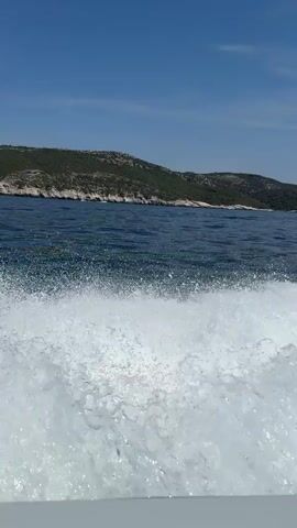 The video shows a boat moving through the water, leaving a wake behind it. The water is blue and there are waves. In the background, there is a coastline with hills. The sky is clear and blue. The camera is mounted on the boat and is pointed backwards. The boat appears to be white and is moving at a good speed.