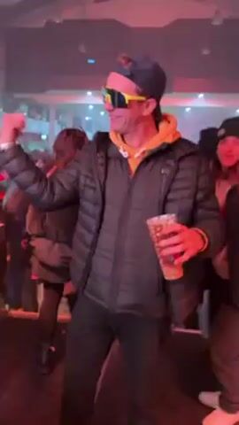 The video shows a crowded party, probably indoors. A man wearing a black puffer jacket, yellow hoodie, a baseball cap, and yellow-tinted sunglasses dances with a drink in his hand. There are other people dressed in costumes dancing behind a DJ table, with the DJ operating the equipment. The scene is lit with red and white lights, creating a party atmosphere. There are balloons and decorations hanging from the ceiling. The crowd is dancing and enjoying themselves.