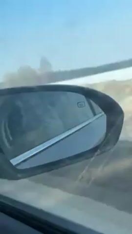 The video shows the side mirror of a car moving down a road. The mirror has a symbol indicating that it is heated. Reflected in the mirror is the blurred image of the driver and passenger. Outside the car window, the landscape is visible, featuring snowy fields, bare trees, and a clear blue sky. The car appears to be traveling on a road with some dirt on the shoulder.