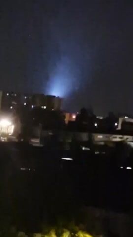 The video is a night-time shot of an urban landscape. In the background, several multi-story buildings are visible, some illuminated with lights. A bright beam of light emanates from one of the buildings, projecting upwards into the dark sky. The foreground is dark, with indistinct shapes that could be trees or other foliage. The overall impression is one of a city at night, with artificial light cutting through the darkness.