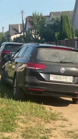 This is a horizontal, medium shot taken from a low angle on a sunny day. In the foreground, the rear of a black Volkswagen Passat station wagon is visible, parked on a dirt road. The license plate reads "0806 PE-5". To the left and slightly behind the Passat is a black SUV, possibly a Ford Kuga, with its license plate reading "2540 DB-4". Both vehicles are dusty. The road is dirt and gravel, with tall green grass growing on the left side. In the background, there are houses with peaked roofs and fences, and several trees, some of which are evergreen. The sky is clear and blue. The video shows a slight movement of the SUV as it appears to be backing up or maneuvering.