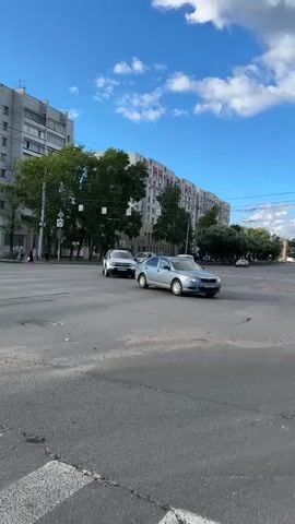 This is a vertical, eye-level video of a street scene in Rubtsovo, Russia. The sky is a vibrant blue with scattered white clouds, suggesting a pleasant day, likely late morning or early afternoon given the lighting. A multi-story apartment building with a long, beige facade and red-capped balconies dominates the background, typical of Soviet-era architecture. Lush green trees line the street in front of the building.

The foreground shows a wide asphalt intersection with clear lane markings and a pedestrian crossing. Several cars are navigating the intersection. A silver Dacia Duster is turning, followed by a silver Skoda Octavia. Another white car, possibly a minivan, is also in motion. Further back, a black SUV is visible. The overall activity is routine urban traffic.

There are no prominent people visible, though a few figures are faintly seen on the sidewalk to the left. A pedestrian crossing sign is clearly visible on the left side of the road. The overall mood is ordinary and uneventful, capturing a slice of everyday life in a Russian city. No discernible text is visible on the cars or any signage besides the pedestrian crossing symbol.