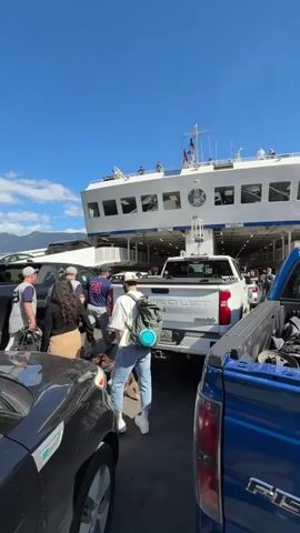 The video shows cars and people boarding a large white ferry under a clear blue sky. Several individuals with backpacks are walking towards the ferry entrance, passing between parked pickup trucks and cars. The ferry is prominently visible, its white exterior contrasting with the bright sky. People can be seen on the upper decks of the ferry. The vehicles are queuing to board, and the overall scene suggests a busy port with passengers and vehicles embarking on a ferry trip.