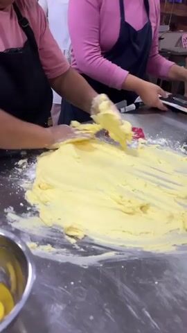 A medium shot shows a woman wearing a pink shirt and a black apron working with a large mound of yellow dough on a stainless steel table. Her hands, covered in gloves, are actively mixing and kneading the dough, which is spread out thinly across the table. Flour is visible on the table and on her hands. In the background, another person in a pink shirt and a dark apron is partially visible, also appearing to be involved in a similar task. Other individuals and various kitchen equipment can be seen in the blurred background, suggesting a commercial kitchen or food preparation area. The lighting is bright and appears to be artificial, illuminating the workspace. The action is dynamic, with the woman's hands in constant motion, indicating she is in the process of preparing a large batch of dough.