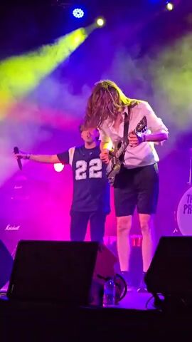 This is a video of a live performance on a stage. There are two men visible. The man on the left is wearing a dark blue jersey with the number 22 on it, a baseball cap, and holding a microphone. He is looking towards the right and gesturing with his left hand. The man on the right is playing an electric guitar and is wearing a white shirt and dark shorts. He has long, dark hair that is falling over his face. The stage is lit with blue and purple lights, and there is a haze or smoke effect. In the background, there are speakers and part of a drum kit with the words "THE PROUD" visible on it. There is a water bottle on the stage in the foreground. The video is shaky and seems to be recorded from the audience.