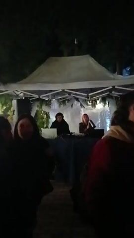 A nighttime video shows two DJs performing under a small, white, tent-like structure. The DJs, one with dark hair and one with reddish hair, are positioned behind a table with laptops and DJ equipment. The tent is decorated with white cloth and greenery. Several people are standing in the foreground, facing the DJs. The overall lighting is dim, suggesting an outdoor event at night. The sound of music is audible in the background.