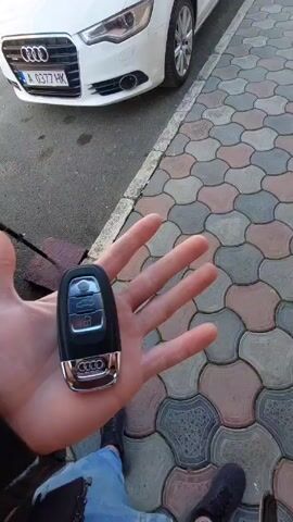 A person holds an Audi car key in their hand in front of a white Audi A6 parked on a street with a patterned sidewalk. The car has a license plate that reads "A 0377 HK". The street is in a town with buildings, trees, and other cars visible in the background. The sky is blue and sunny. The person is wearing jeans and black shoes. The camera angle changes slightly throughout the video to show different aspects of the scene, but the primary focus remains on the car and the key.