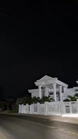 At night, a stately white house with pillars is visible behind a white fence along a black road. A single motorcyclist passes on the road in front of the house. The sky is completely dark. The house is illuminated with lights that highlight the architectural details of the building.