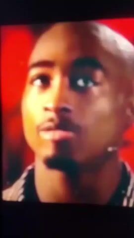The video is a close-up shot of a Black man speaking. In the first few frames, the man is bald and has a mole on his cheek. He is wearing a dark and striped garment. The background is blurred and appears to be red. In the subsequent frames, the man is wearing a white bandana and a knitted vest over a light-colored shirt. His skin appears lighter, and he has a earring in his left ear. The background is a solid bright blue. The man's mouth is slightly open as if he is speaking. The video is grainy and low resolution.