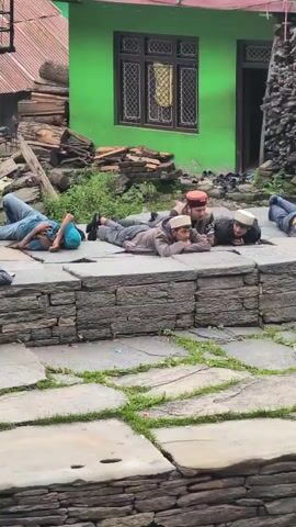 The video shows a group of people lying down on a stone platform in what appears to be a village setting. The individuals are dressed in casual clothing, and some are wearing traditional caps. The background features a green building, stacks of wood, and other structures typical of a rural environment. Some people are looking towards the camera, while others appear to be resting or sleeping. The overall atmosphere is relaxed, with a sense of community. The scene is captured from a slightly elevated angle, offering a clear view of the platform and the people on it.