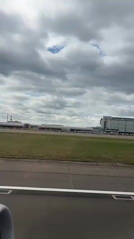 The video shows an aerial view of an airport, likely taken from an airplane window. In the foreground, there is a grassy field and a tarmac. In the background, a large, modern airport terminal building is visible, with numerous parked airplanes and ground support vehicles. The British Airways logo is prominently displayed on the side of the building. The sky is cloudy, suggesting overcast weather conditions. The camera angle is slightly tilted, giving a dynamic perspective of the airport and its operations.