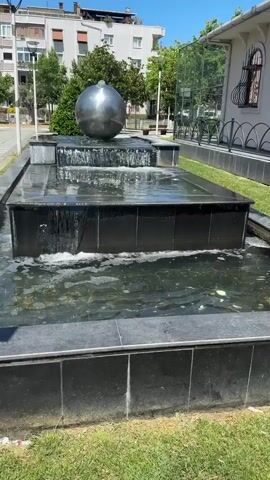 The video pans across a multi-tiered fountain with a large, spherical metallic sculpture at its highest level. Water cascades down the dark, reflective tiers of the fountain. The fountain is surrounded by green grass and a paved walkway. In the background, there is a building with arched windows and ornate metalwork, as well as trees and a taller apartment building. The sky is bright blue and the scene appears to be outdoors on a sunny day. There are no people visible in the video.