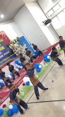 This video captures a group of young boys, likely performing a dance routine on a stage. They are all dressed in matching dark blue outfits with ornate patterned sashes and dark sunglasses, giving them a coordinated and somewhat stylish appearance. The boys are spread across a red carpeted stage, with a backdrop featuring a banner in yellow, blue, and red text, indicating an event or celebration. White and blue balloons are strategically placed around the stage, adding to the festive atmosphere.

The boys appear to be in motion, performing synchronized dance steps, as suggested by their varied poses and the dynamic composition of the video. Their expressions are difficult to discern due to the sunglasses, but the overall energy of the performance seems upbeat and engaging. The setting appears to be an indoor venue, possibly a school hall or community center, given the large windows and the lighting. The event is likely a school performance, a cultural presentation, or a talent show, given the formal attire and staged presentation.

The visible text on the banner is in Indonesian. While not fully legible, parts of it can be deciphered, mentioning "ANAK" (child), "KREATIFITAS" (creativity), "PERPISAHAN" (farewell), "KAB. PIDIE" (Pidie Regency), and "SELALU MENGGENGGAM SEJARAH" (always holding history). This suggests the event might be a graduation or end-of-year celebration for children in the Pidie Regency of Indonesia, with an emphasis on creativity and heritage. The time of day is not explicitly indicated, but the bright indoor lighting suggests it could be during daylight hours. The overall mood is celebratory and spirited, highlighting the children's performance and the significance of the occasion.