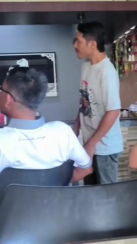 The video shows an indoor scene, possibly a cafe or barbershop, with several people present. In the foreground, a person in a white polo shirt with a gray collar is seated, facing away from the camera, with their back and the side of their head visible. They are wearing glasses. To the right, another person in a gray t-shirt is standing and looking at the seated person. Behind them, a mirror hangs on the wall, reflecting a portion of the room. Further back, shelves are visible with various items, including bottles and containers. On the left side of the frame, another person in a beige hooded jacket is partially visible, facing to the left. A television screen is mounted high on the wall, displaying a soccer match with players in white and orange uniforms on a green field. The environment appears somewhat cluttered, with visible signage and structural elements. The lighting suggests an indoor setting during daytime.