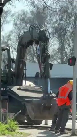 The video shows a construction scene in what appears to be a European urban area. Two construction workers wearing yellow hard hats and orange safety vests are working on the side of the road near a pedestrian crosswalk signal displaying a red walking person. One of the workers is holding a broom, and the other has a long-handled tool, possibly a shovel or scraper. Behind them, a large construction vehicle, seemingly a backhoe, sits idle with its arm raised. The area around the workers has been disturbed, with dirt and debris visible. The background includes trees without leaves, a white building, and possibly other traffic. The day appears sunny.