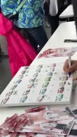 The video shows a person signing a poster of a car team calendar with a blue marker. The poster is displayed on a white table alongside a stack of cards with the text “THEO SEVAHAUS” printed on them.  The person is signing a square with a drawing of a race car on the poster. To the left, another person in a blue patterned shirt stands holding a bright pink bag. Several other individuals can be seen standing around the table, likely waiting in line to receive autographs.