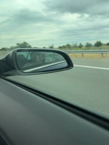 The video captures a scene from inside a moving vehicle, looking out the side window and into the side-view mirror. The perspective suggests the video is being recorded from the passenger seat. The side-view mirror reflects a person, likely the person recording, holding a smartphone. The background visible through the car window shows a highway with a guardrail and dry grass on the side. The sky is cloudy. The vehicle appears to be on a paved road, possibly a highway, indicated by the lane markers. The overall impression is that of a journey or travel.
