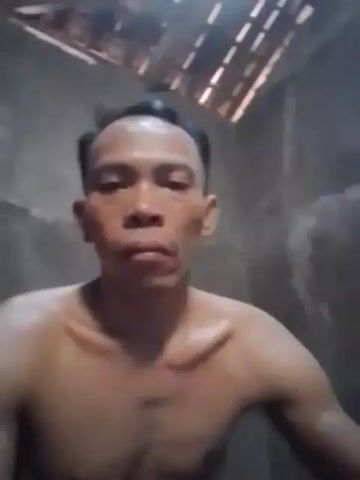 A man's bare shoulders and upper chest are visible from the chest up. He has dark hair, and his skin is brown. He is looking directly at the camera with a neutral expression. The background appears to be a room with rough, gray walls. Above the man, there are wooden beams. The lighting is dim and appears to be coming from overhead.
