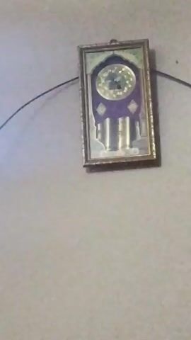 The video shows a framed picture hanging on a light grey wall. The frame is gold and the picture appears to be of a building with a clock in the background. A black wire hangs from the top left corner of the frame and continues out of view. The image is slightly out of focus.
