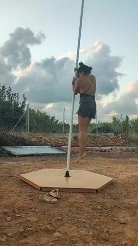 A person is performing a pole dance routine outdoors. They are wearing a black top and denim shorts. The pole is set up on a hexagonal base on a dirt ground. In the background, there is a fence, some trees, and a cloudy sky. There are also a pair of shoes on the ground near the base of the pole. The person is shown moving around the pole, spinning and holding different poses.
