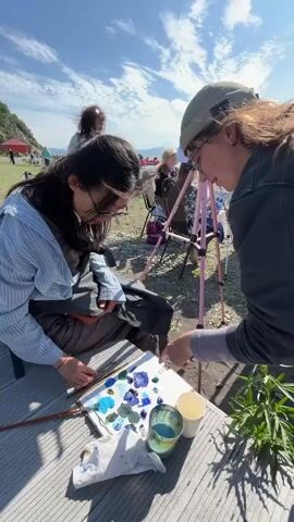 A group of people are gathered outdoors on a beach with a body of water in the background. Two people in the foreground are focused on a palette of blue paints. One person, wearing a light blue striped shirt and glasses, is holding a paintbrush and looking down at the palette. The other person, wearing a dark jacket and a beige cap, is pointing at the palette, seemingly instructing the first person. Several other individuals are visible further back, some appearing to be painting or observing. The sky is blue with scattered white clouds, and the sun is shining. The ground is covered with pebbles, characteristic of a beach.