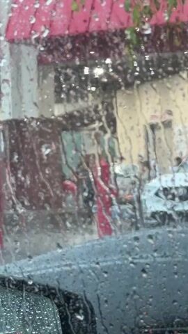 From inside a car, rain is visible on the windshield. Through the rain-streaked glass, there is a view of a street scene with buildings and cars. One building has a red awning. People are partially visible, moving on the sidewalk. The windshield wipers of the car are not in operation. The scene suggests a rainy day in a city or town.