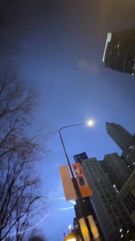 The video is a low-angle shot taken at night in an urban environment. Initially, lightning is visible in the sky, followed by a view of the night sky obscured by clouds. A streetlight is visible in the foreground, with two banners attached to it. The banners appear to promote "Jazz" and "Rhythms in Color." The surrounding buildings are tall and typical of a city skyline. Bare tree branches are also visible in the lower left corner of the frame. The camera appears to be held by someone looking upward.