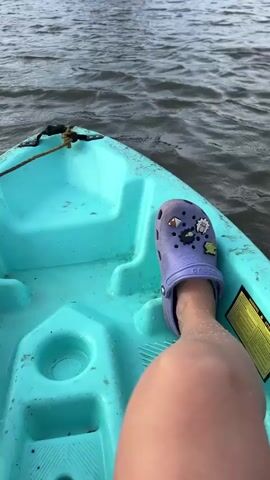 The video opens with a close-up of a person's leg wearing a purple Croc shoe with charms on it, resting on the edge of a teal-colored kayak. The kayak is on dark, rippling water. The camera then pans out to reveal a wider view of the kayak moving across a body of water. In the distance, a lush green shoreline with trees is visible under a cloudy sky. A solitary white bird, likely an egret, stands in the shallow water near the shore. The video continues to show the bird as the kayak approaches, and the camera remains focused on the tranquil scene of the bird and the natural landscape.