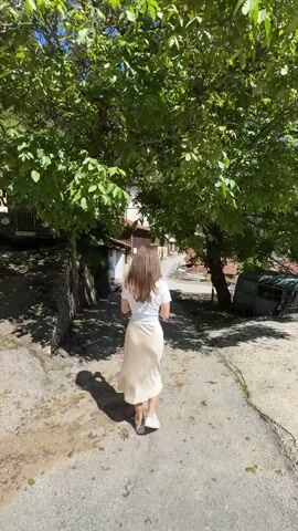 A young woman with long brown hair walks away from the camera down a dirt and gravel path. She is wearing a white t-shirt and a light-colored satin skirt. Her shadow is visible on the path behind her. The path is lined with trees with green leaves and there are some buildings visible in the background. The scene appears to be a rural village or a countryside setting. The lighting suggests it is daytime, possibly morning or afternoon.