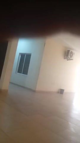 The media file captures a dimly lit interior space, likely a hallway or an empty room, with tiled flooring. A window with vertical blinds is visible on the left wall, set against a lighter-colored wall. The room appears to be furnished with minimal items, including a dark-colored object on the floor in the distance, possibly a bin. An air conditioning unit is mounted on the right wall. The scene is dominated by soft, diffused light entering from an unseen source, suggesting either natural light from a window or a light fixture. The overall impression is one of stillness and emptiness. The location is contextualized as Arawa, Nigeria. There is no discernible activity taking place, and no people are visible. The time of day and weather are not evident from the visual information. The image quality is blurry, with a significant portion of the foreground obscured by a dark, out-of-focus area that appears to be a finger or hand. No text is visible within the frame.