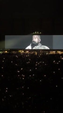 A distant, high-angle shot showcases a dark concert venue. A large screen displays a profile view of Kanye West, clad in a gray jacket and sporting a beard. His head is shaved, and he is holding a microphone close to his mouth. Below the screen, a massive crowd is visible, their faces obscured by darkness, save for the small, glowing lights of cell phones held aloft. The atmosphere is dimly lit, with a few faint lights visible in the far background.