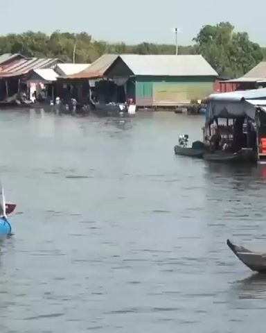The video shows a body of water with several floating structures in the background, appearing to be houses. In the foreground, a wooden boat is moving from left to right across the screen. There are two people in the boat. The person in the front is wearing a wide-brimmed hat and appears to be looking forward. The person in the back is holding an oar and appears to be rowing the boat. The water is calm and appears to be reflecting the sky and the structures. The overall scene is peaceful and suggests a rural or village setting.