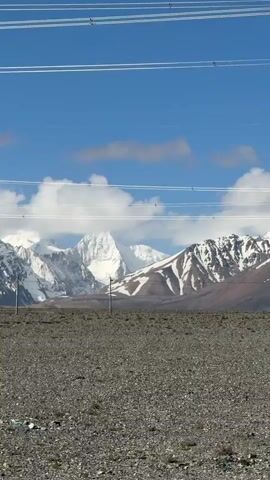 The video displays a vast, barren landscape under a clear blue sky with scattered white clouds. In the background, majestic snow-capped mountains stretch across the horizon, their peaks glistening in the sunlight. The foreground consists of rough, rocky, and gravelly terrain, suggesting an arid or high-altitude environment.

Initially, only the distant mountains and the rocky ground are visible, with several parallel power lines traversing the upper portion of the frame. As the camera slowly pans to the left, a large, metal lattice power transmission tower (pylon) comes into view, standing tall in the midground. The power lines extend from this tower and across the scene, cutting across the sky and mountains. The scene conveys a sense of remoteness and the impressive scale of both nature and human infrastructure in Gyirong County, China. The day is bright and sunny. No people or visible text are present.