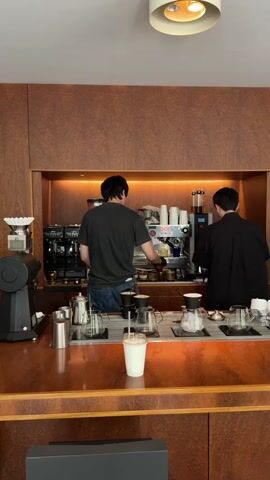 Two young men are working behind a coffee bar. The first is on the left, wearing a gray t-shirt and jeans. He is operating a complex espresso machine, wearing a glove on his right hand as he prepares the espresso. The second man, on the right, wears a black button-down shirt and is preparing coffee using a pour-over method.  Various pieces of coffee-making equipment such as a grinder, kettles, pour-over sets, and an espresso machine are arrayed on the countertop. A drink in a disposable cup sits on the counter in front.  The back wall is wood paneling, with overhead lighting highlighting the work area.  The countertop appears to be varnished wood with a distinctive patterned edge.