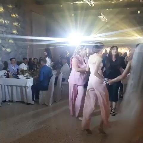 The video shows a group of people dancing at a formal event in a dimly lit room with spotlights. In the foreground, two women are dancing, one in a pink jumpsuit and the other in a light pink dress. They are holding hands and moving their feet. Behind them, other people are visible, some sitting at tables and others dancing. The room appears to be a banquet hall or reception area with white tablecloths on the tables and chairs arranged around them. The lighting is dramatic, with bright spotlights creating beams of light that crisscross the room. The overall atmosphere seems festive and celebratory.