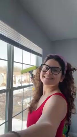 A woman in a red top and glasses stands in front of a window with white blinds and a grid pattern. She has curly hair and a pink headband. She is using a green and yellow squeegee to clean the window. Outside the window, there is snow on the ground and houses in the background. The room has gray walls. The woman turns her head and smiles towards the camera briefly before continuing to clean the window. The sound of a baby cooing can be heard.